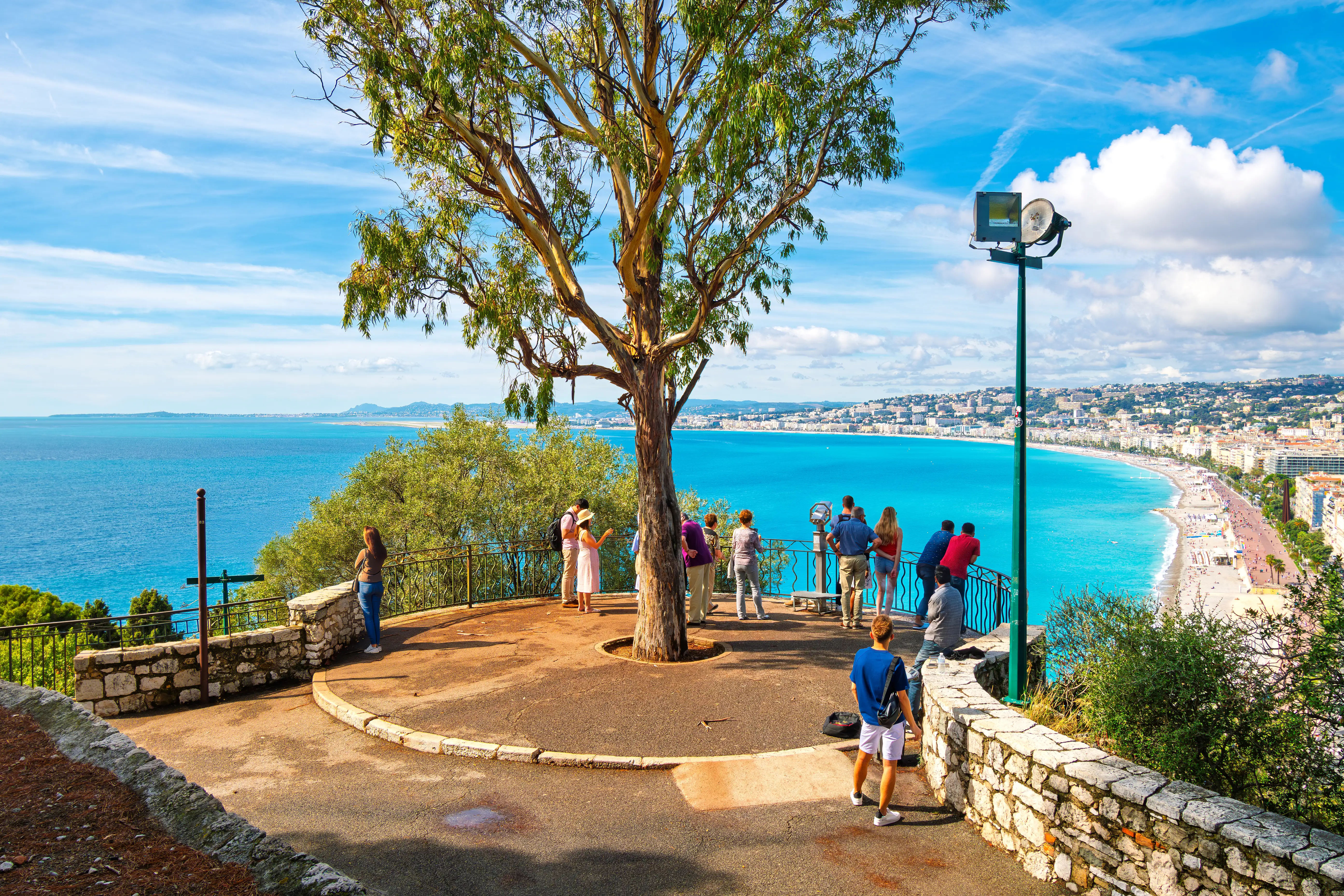 Tourists enjoying a sunny day on Castle Hill, overlooking the turquoise Mediterranean Sea, pebble beach, and the cityscape of Nice on the French Riviera.
