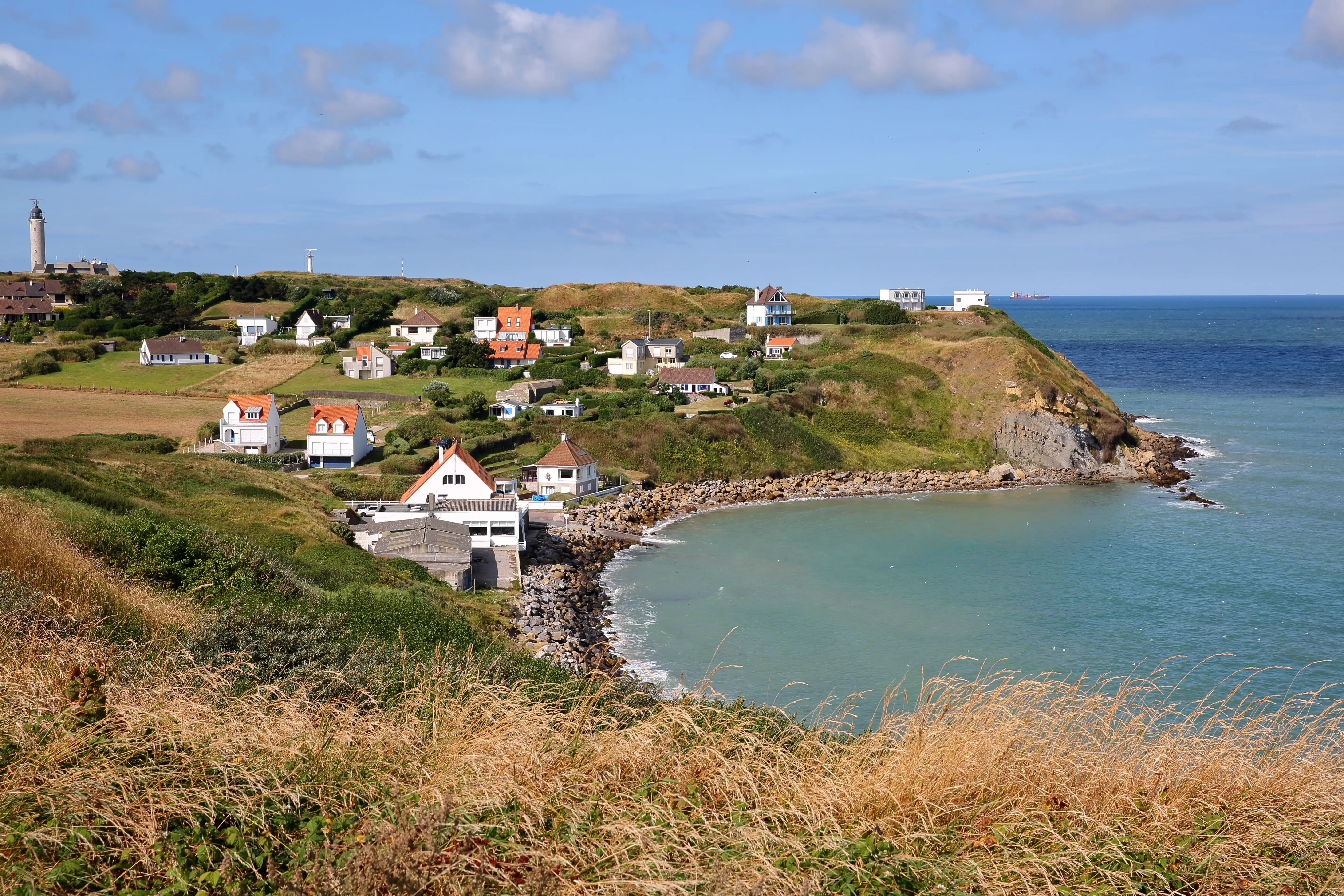 A coastal headland with white houses dotted around the hill above the shore, with a lighthouse to the left. Cap Gris-Nez, France.
