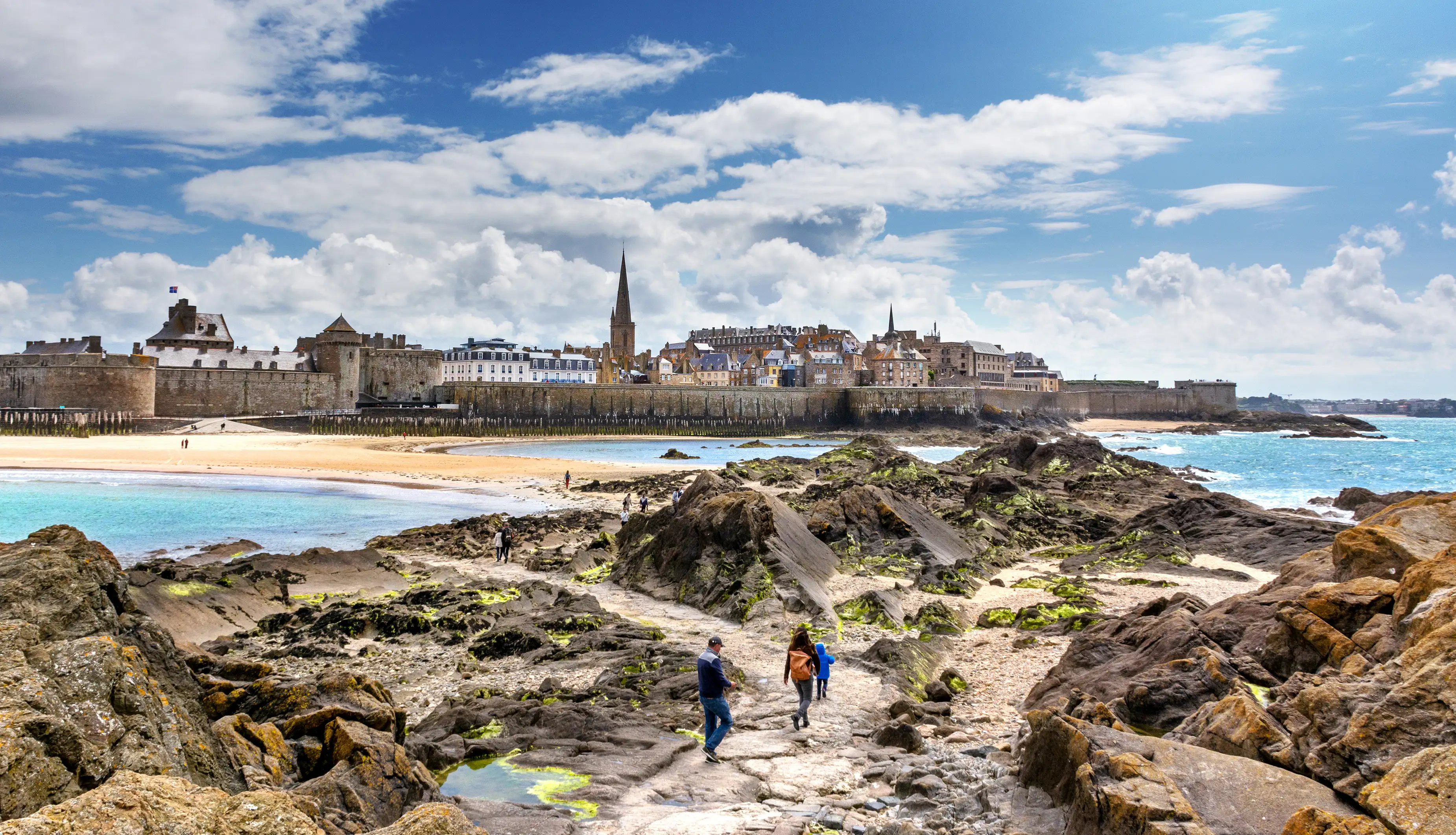 People walking over rocks near a fortified coastal town. Saint-Malo, France