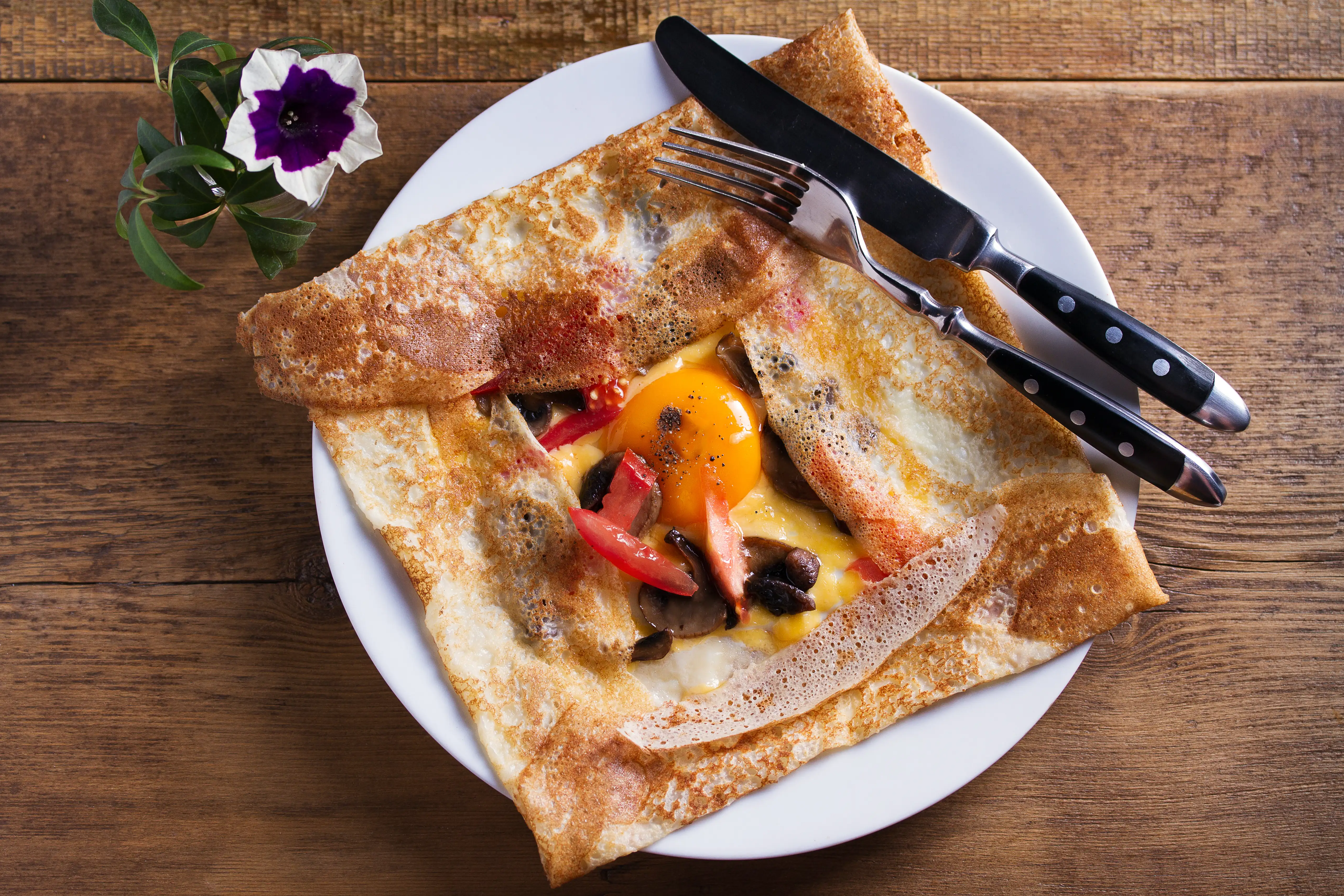 A crêpe on a plate with egg, tomatoes and mushrooms, and a knife and fork