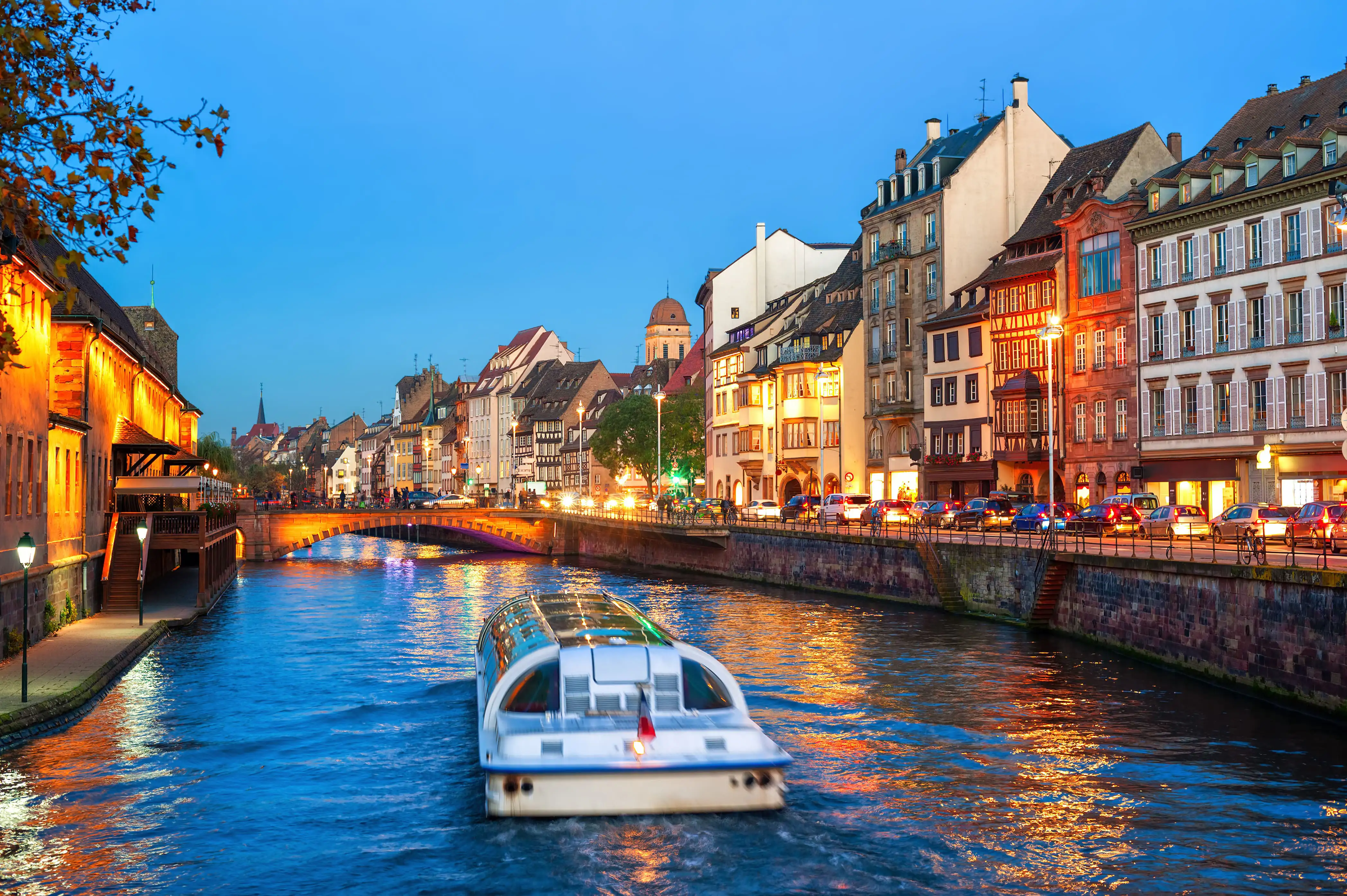 Boat cruising along Strasbourg canal at dusk, surrounded by warm lights reflecting on the water.