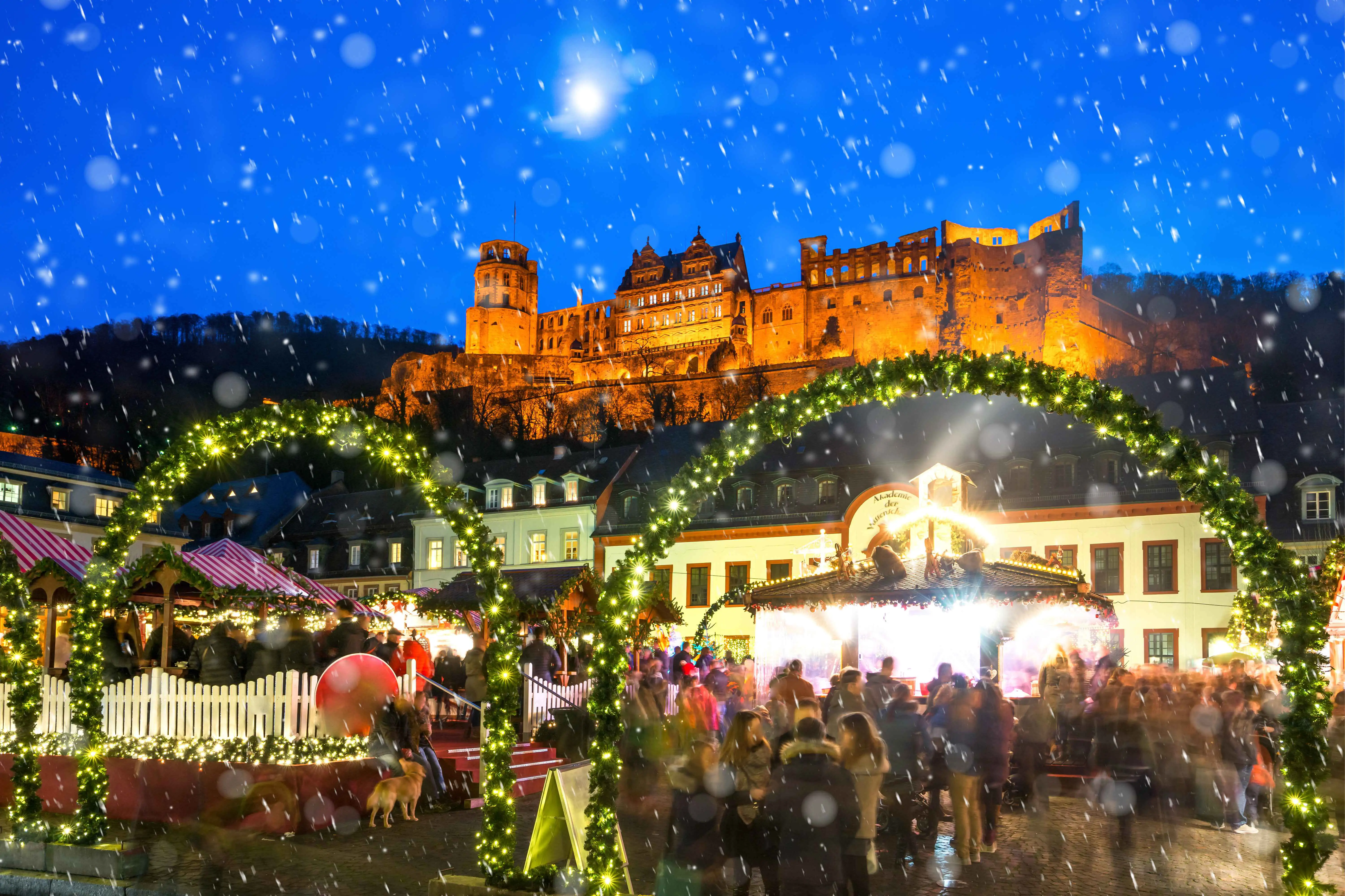 Christmas market scene in a square with snow falling, and Heidelberg castle illuminated above
