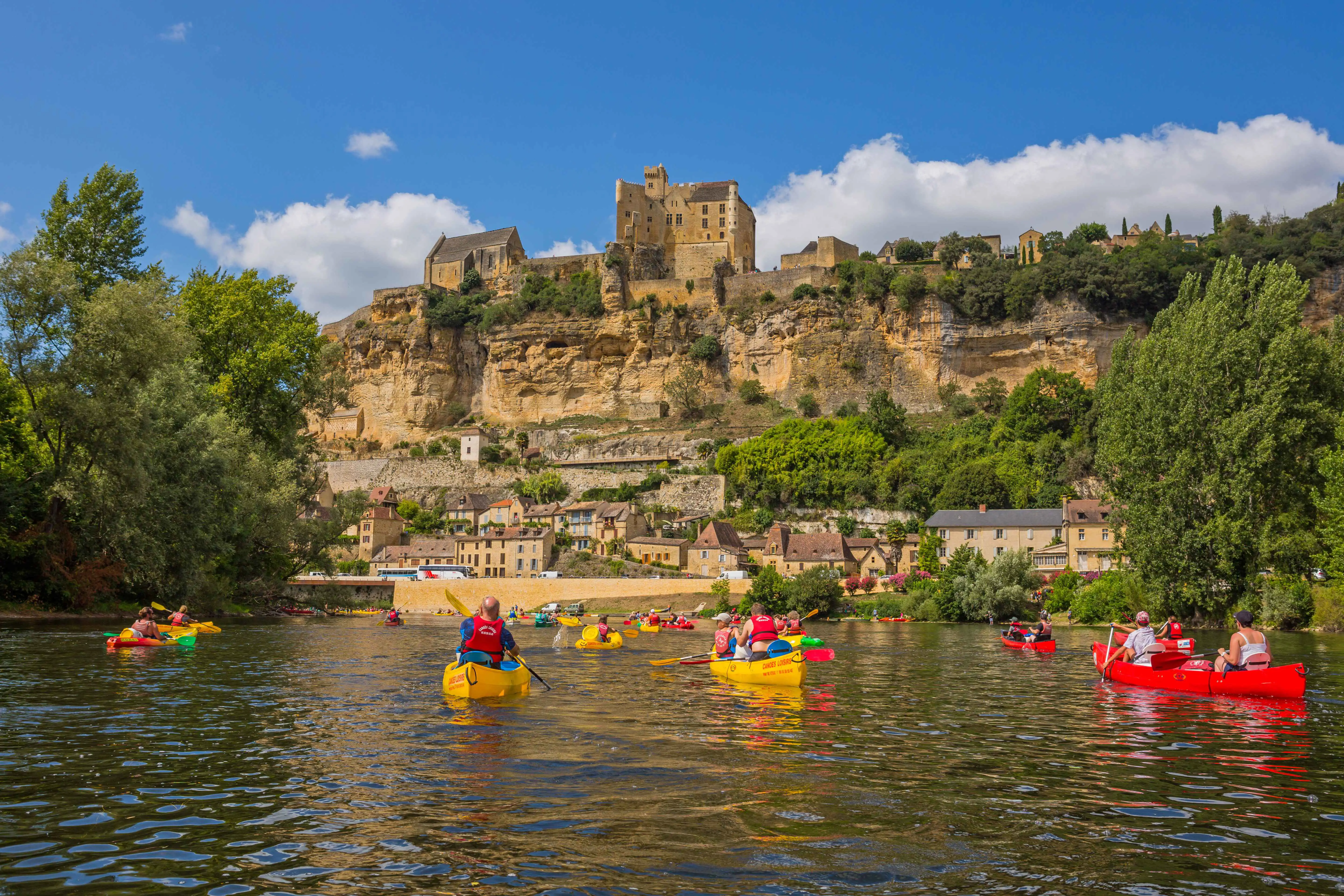 People canoeing on the Dordogne River, surrounded by lush greenery, with a historic building visible ahead
