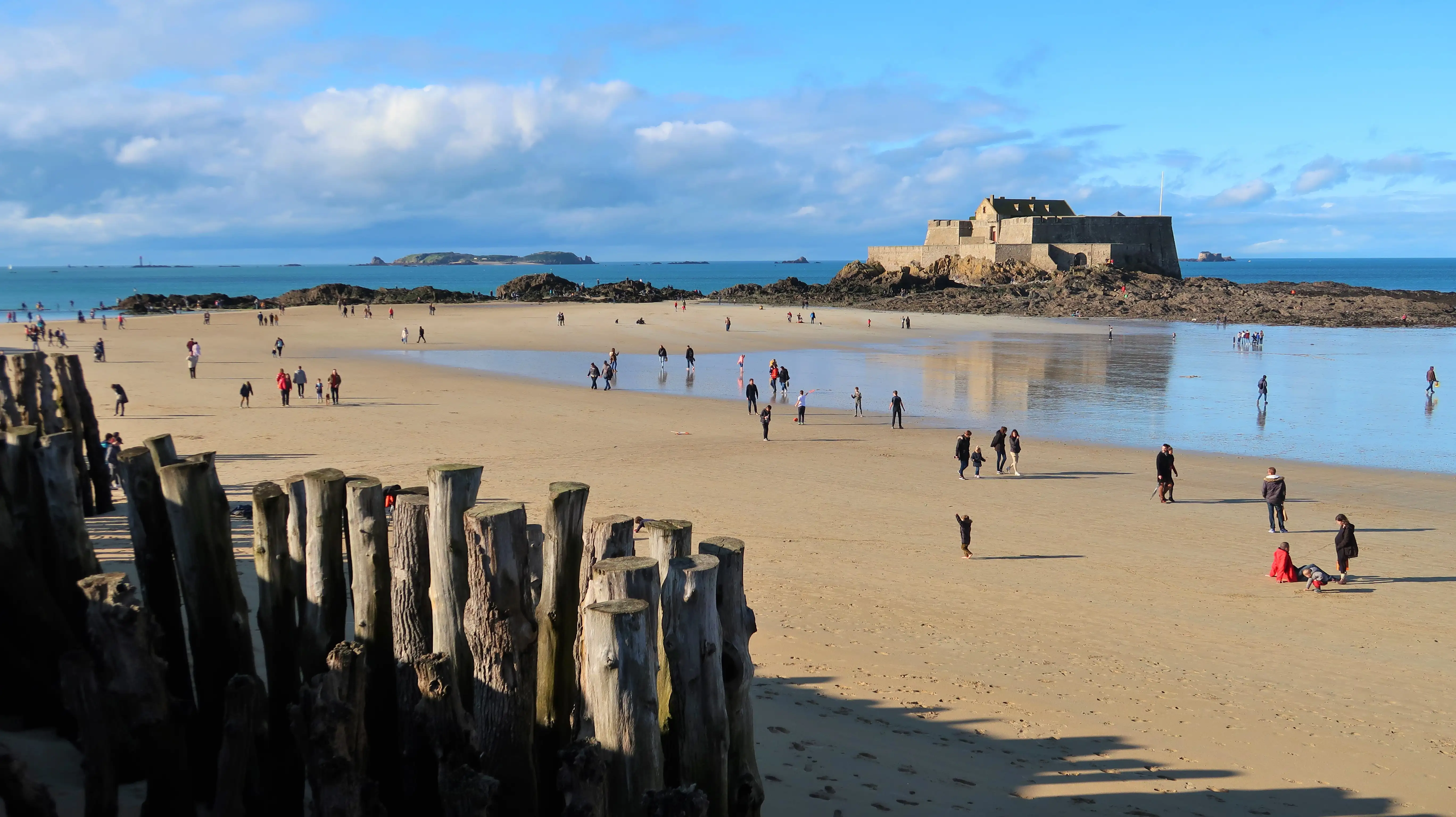 People walking on a wide sandy beach on a sunny winter’s day, with a fort in the distance