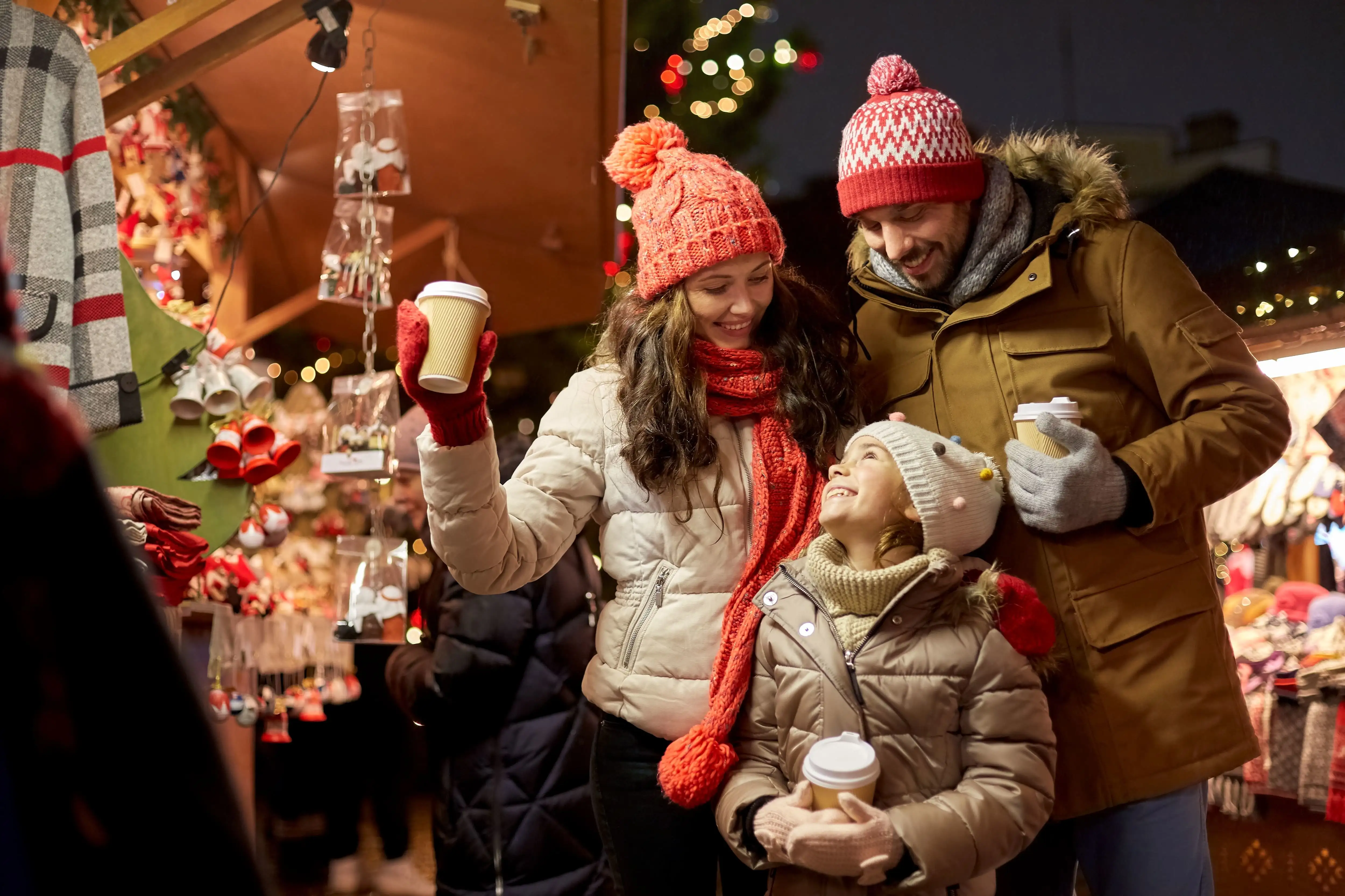 Smiling mother, father, and young daughter holding takeaway drinks while enjoying a Christmas market.