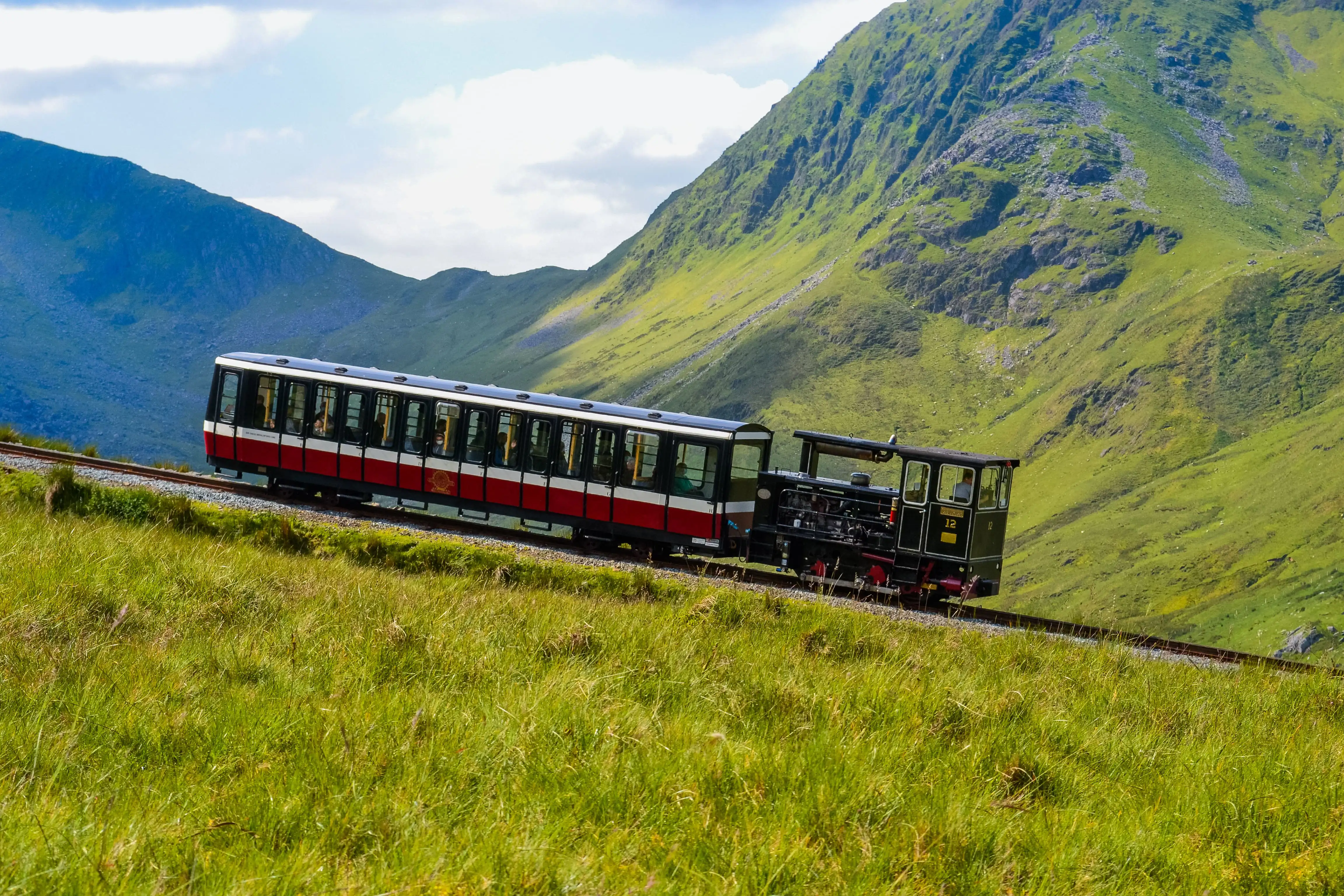 De Snowdon Mountain Railway loopt omhoog over de berg en is omgeven door groen gras, bergtoppen op de achtergrond en een bewolkte lucht.