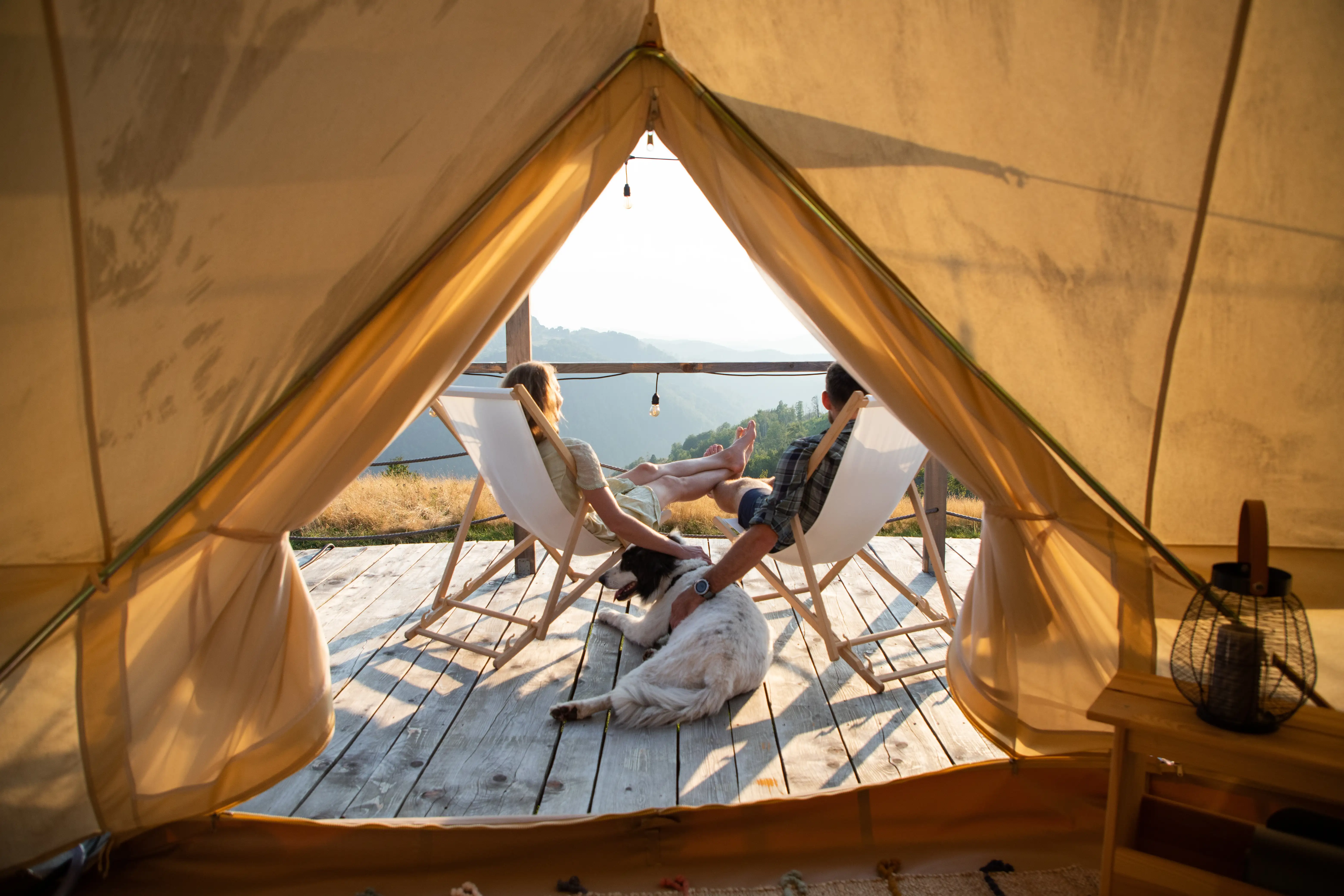 A couple sitting on the decking outside their glamping tent, legs crossed over each other, with their dog resting on the floor beside them.