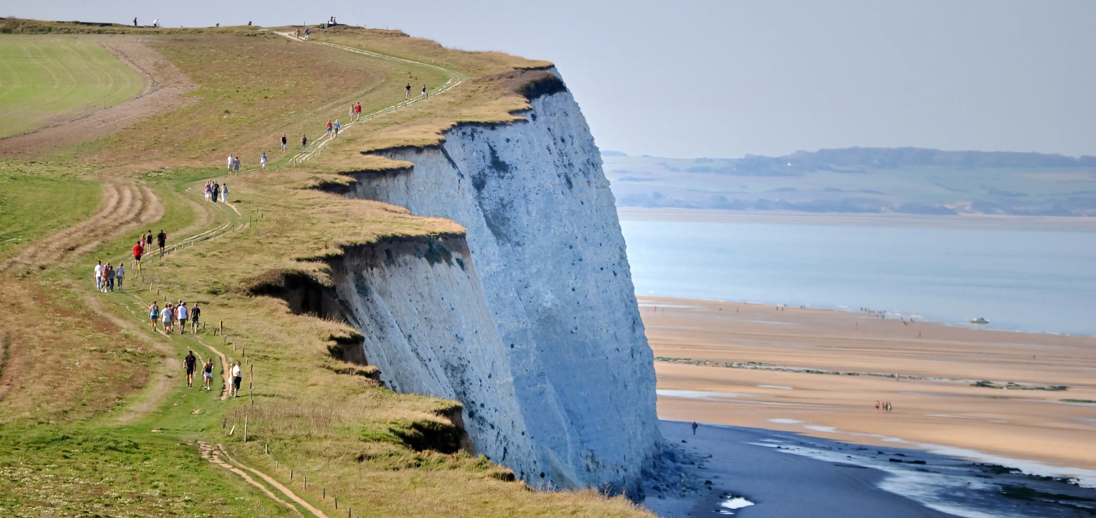 People walking along a footpath on a high grassy headland above white cliffs and a sandy beach