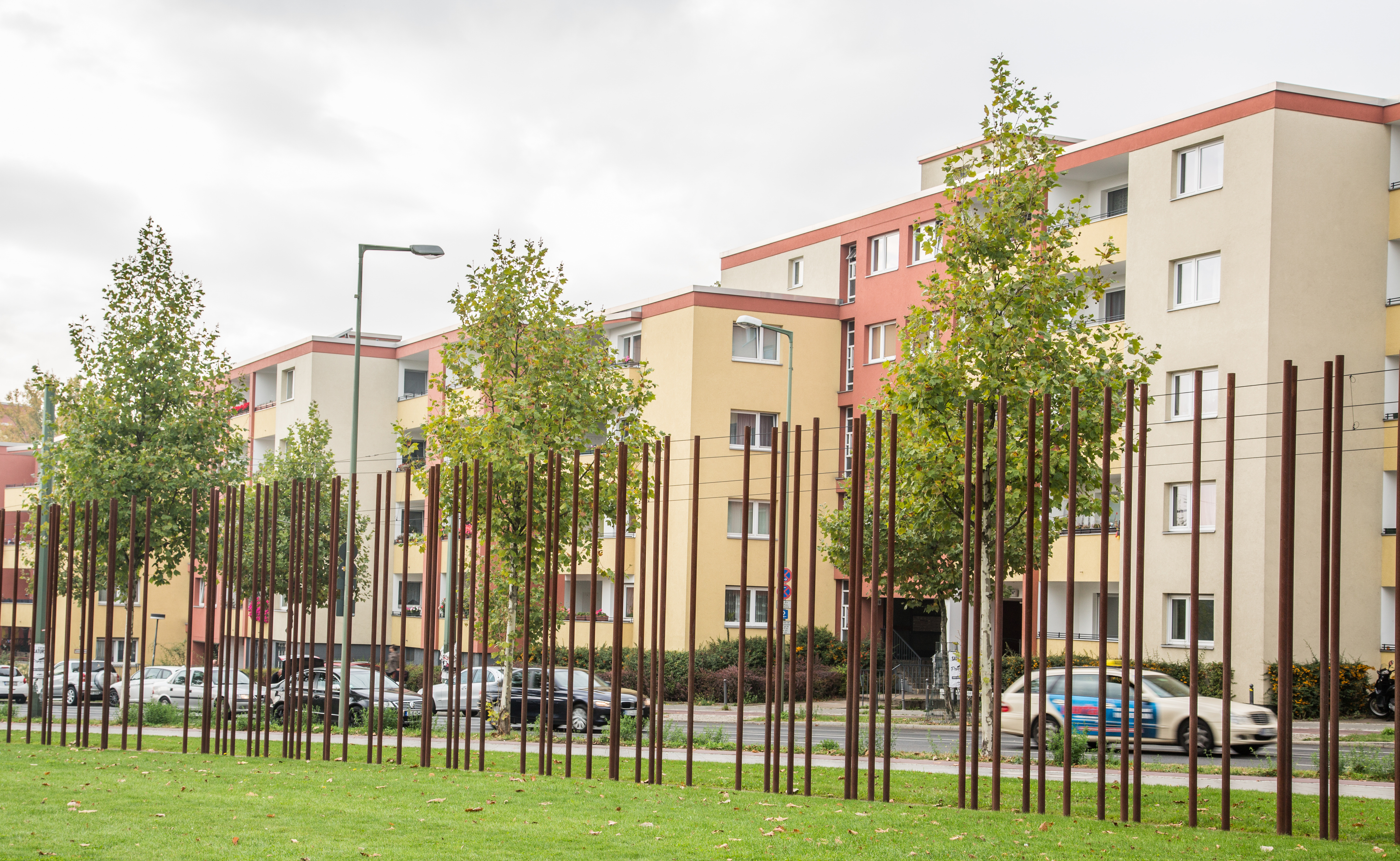 Upright red iron poles mark the path of the Berlin Wall. They stand a green space with a road and homes in the background.