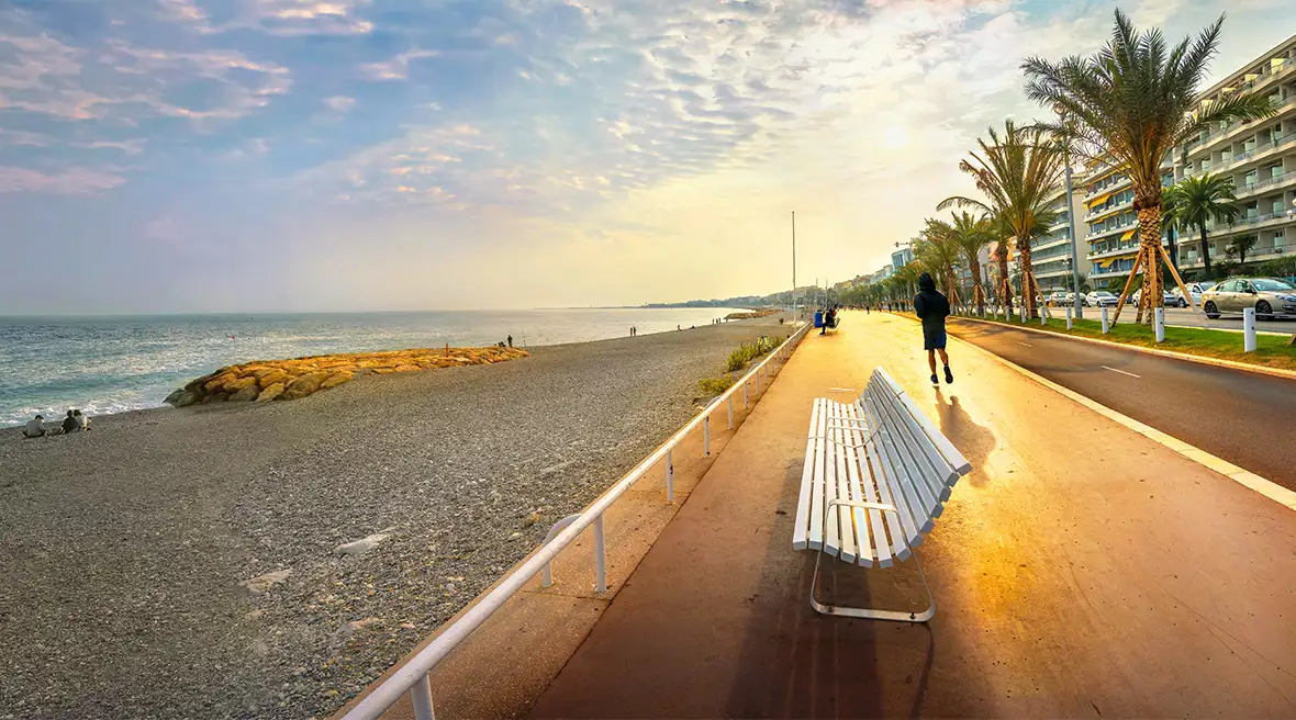 View of the Promenade des Anglais in Nice as the sun is setting, a wide esplanade with a beach to one side and palm trees and hotel to the other.