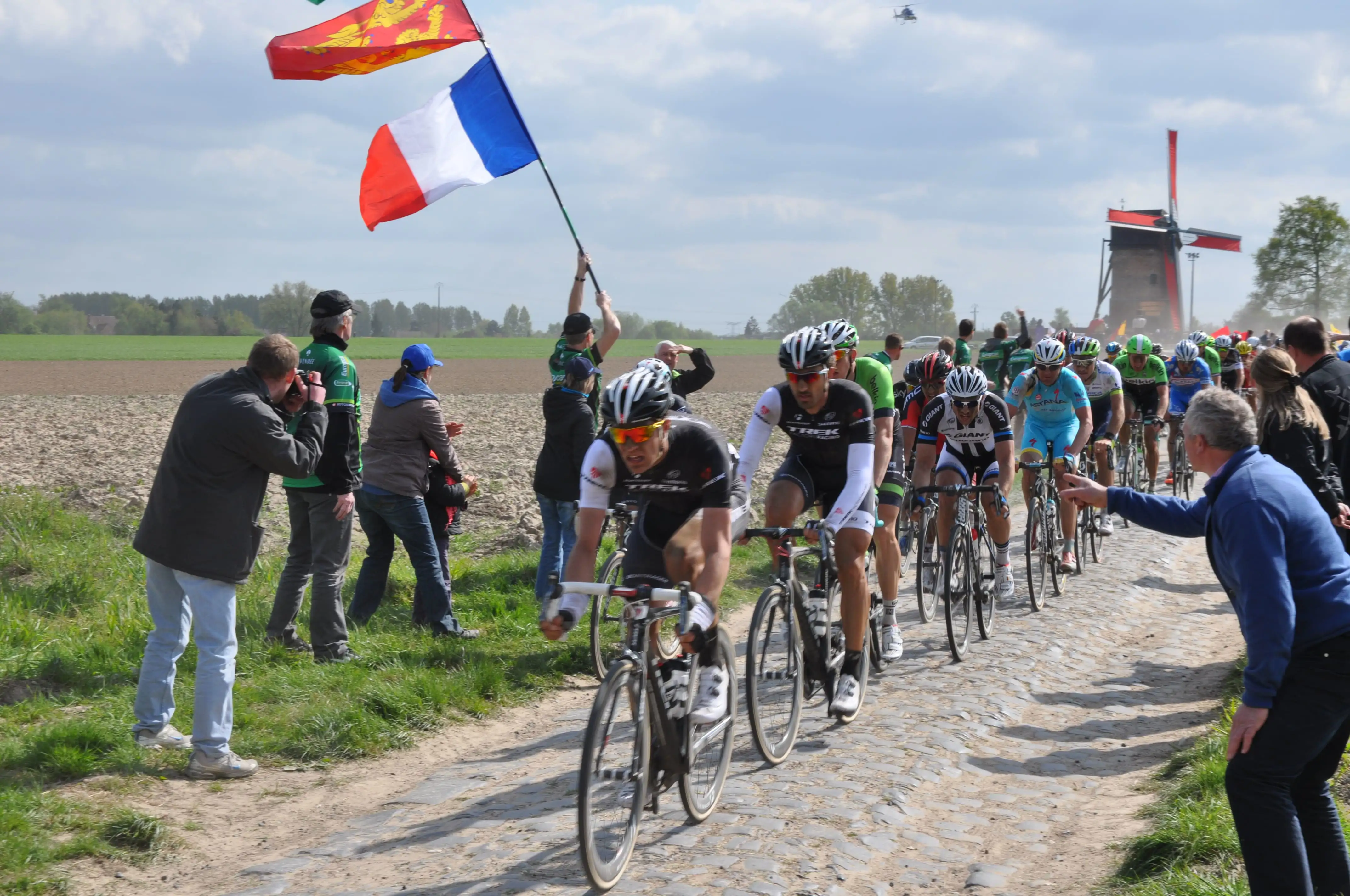 Cyclists ridding over cobbles with supporters encouraging them, holding flags and taking photographs, Paris-Roubaix.