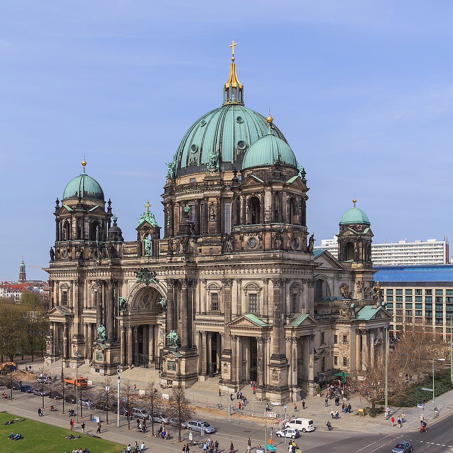 A view of the Berlin Cathedral with its green dome, elaborate columns and intricate carvings. Below the cathedral is a road with moving cars and pedestrians. A clear blue sky is above.