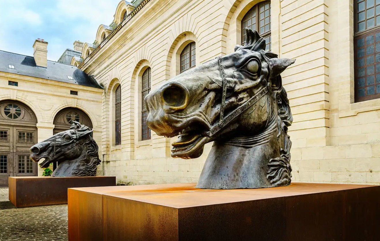 Two large horse heads cast in bronze standing on plinths outside a grand looking building with large glass windows